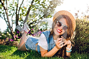 Pretty girl with long curly hair in hat is lying on grass in summer park. on sunlight. She wears jeans jerkin, short