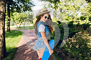 Pretty girl with long curly hair in hat and sunglasses is walking with skateboard in summer park. She wears jeans jerkin