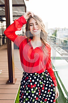 Pretty girl with long hair is posing on terrace. She wears red shirt and black skirt. She is smiling to the camera