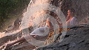 Pretty girl with long hair in a white dress lies with a guy among the rocks under a waterfall and strokes his face