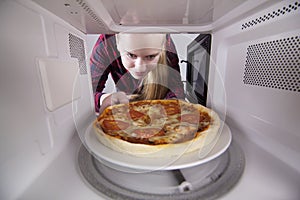 Pretty girl looking in microwave is holding plate with pizza