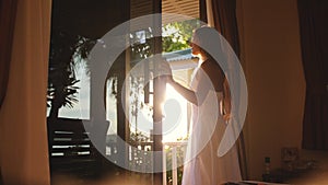 Pretty girl opens the door in the early morning during sunrise with lens flare effects and look out to the shining sun