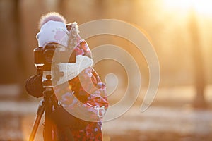 Pretty girl photographer taking pictures on camera using tripod, sunset light, winter, copyspace