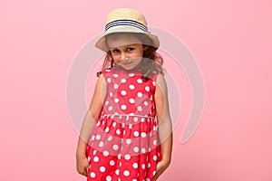 Pretty girl in a pink summer dress with white polka dots and a straw hat, looking shyly at the camera, posing against a pink wall