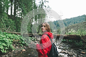 Pretty girl in a red raincoat and with a backpack on her back stands on a stream in the wild nature of mountain landscape and