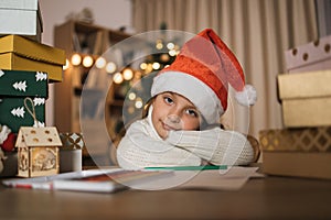 Pretty girl in Santa hat writing letter and joyfully lying at table near the pile of presents