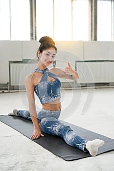 Pretty girl sits in longitudinal twine on fitness mat in industrial room and shows her big finger up