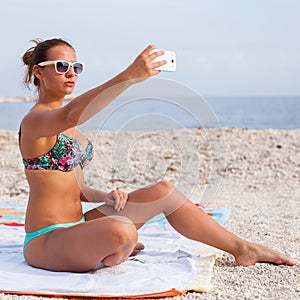 Pretty girl sitting on the beach and taking selfie by her phone.