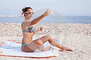 Pretty girl sitting on the beach and taking selfie by her phone.