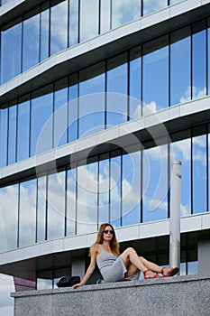 Pretty girl sitting on marble surface with bent knee. Reflection of the sky in windows