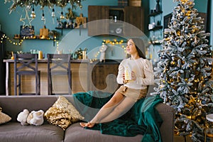 Pretty girl sitting on the sofa near the Christmas tree and holding a toy in her hands a glowing cozy house in anticipation of a