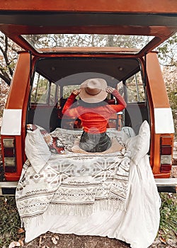 Pretty girl is sitting in the trunk of a red retro minivan. View from the back. Blanket and pillows in Scandinavian style on her
