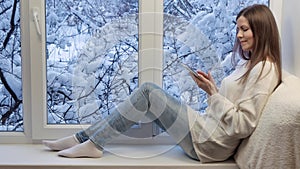 Pretty girl sitting on the windowsill, looking the window and using tablet computer. Winter outside.