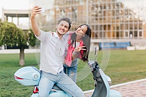 Pretty girl with straight long hair posing with kissing face expression near scooter. Laughing man in stylish white