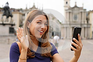 Pretty girl video calling and greets with mobile phone during her travel in Europe. Young traveler woman on landmark city square