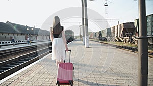 Pretty girl walks with suitcase on platform and waving her hand to the train