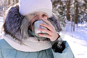 pretty girl in warm clothes drinks tea from thermos in winter forest. Outdoor recreation on frosty sunny day.