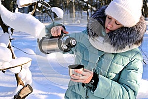 pretty girl in warm clothes pours tea into mug from thermos in winter forest. Outdoor recreation on frosty sunny day.