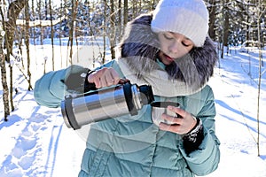 pretty girl in warm clothes pours tea into mug from thermos in winter forest. Outdoor recreation on frosty sunny day.