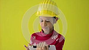 Pretty girl wearing hardhat and red sweater using digital tablet isolated on yellow background. Childhood inspiration.