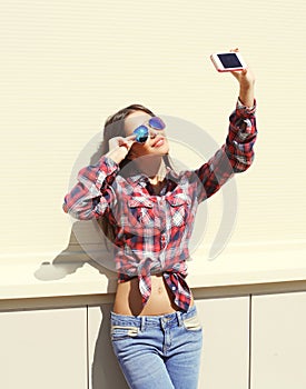 Pretty girl wearing a sunglasses and checkered shirt makes self-portrait on the smartphone outdoors