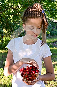 pretty girl in white T-shirt is standing in garden and holding glass vase with red ripe cherries. Concept of gardening