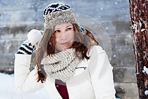 Pretty and fierce girl in winter clothing, scarf and mittens throws a snow ball in front of a wooden barn