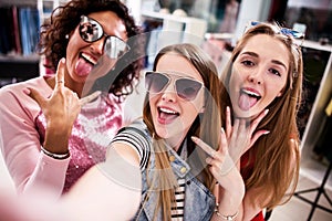 Pretty girls wearing sunglasses fooling around taking selfie showing tongue and horn gestures in clothing shop photo