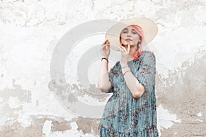 Pretty gorgeous young woman with chic pink hair in fashionable white  hat in summer stylish blue floral dress posing near wall on