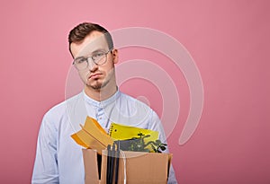 A pretty guy in a sky blue shirt and computer glasses stands on a pink background with a cardboard box with different things in