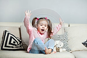 Pretty happy little girl in casual wearing sitting on sofa with toy dog and smiling.