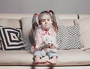 Pretty happy little girl in casual wearing sitting on sofa with toy dog and smiling.