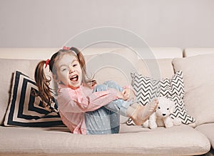 Pretty happy little girl in casual wearing sitting on sofa with toy dog and smiling.