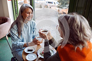 Pretty happy mature women companions hold cups of hot drinks at small table in cozy cafe