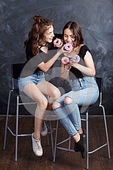 Pretty happy teenage girls with donuts  siting on chairs and having fun. Portrait of joyful smiling girls with donuts on black