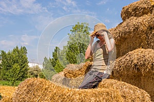 Pretty happy woman with hat sitting on straw bales covering her eyes with her hands.
