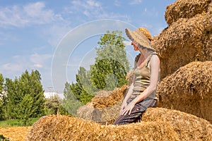 Pretty happy woman with hat sitting on straw bales and getting some rest.