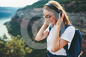 Pretty happy young tourist woman with backpack and  hipster glasses smiling listens favorite music with headphones enjoying