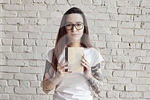 Pretty hipster tattooed woman poses in white t-shirt, isolated on white brick wall, Hold a book in hand, space for design layout.