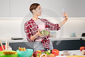 Pretty housewife taking selfie or making a video call using her smartphone while cooking fresh salad wearing a plaid