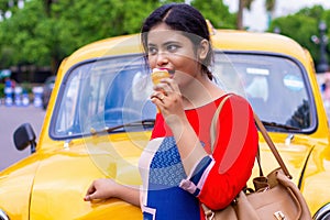 Pretty Indian/asian young girl eating Ice Cream in cone, standing near ice cream shop or taxi