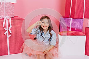 Pretty joyful young girl in tulle skirt sitting suround colorful giftboxes on pink background. Lovely sweet moments of