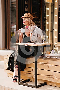 Pretty lady in long dress and black sandals enjoying lunch in outdoor cafe and looking away. Fascinating blonde girl in