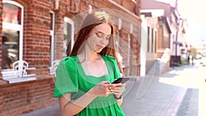 Pretty laughing young woman woman wearing green dress using browsing phone standing at city street in sunny summer day.