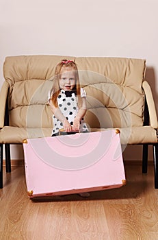 Pretty little blonde girl drags big pink suitcase near sofa