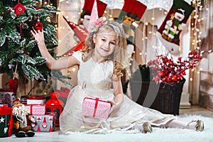 Pretty little blonde girl smiling with a flower hair clip and beige dress with present decorates a Christmas tree The concept of C