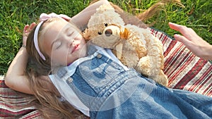 Pretty little child girl with closed eyes laying down with her teddy bear toy on blanket on green grass in summer taking a nap.
