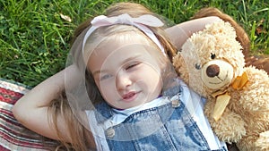 Pretty little child girl laying down with her teddy bear toy on blanket on green grass in summer smiling happily.