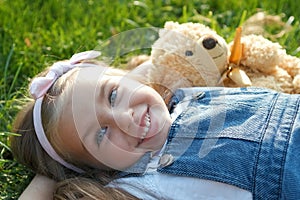 Pretty little child girl laying down with her teddy bear toy on blanket on green grass in summer smiling happily