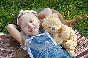 Pretty little child girl laying down with her teddy bear toy on blanket on green grass in summer smiling happily